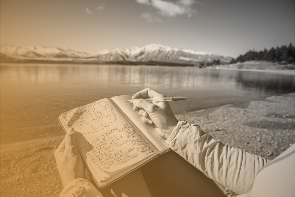Una persona scrive nel suo diario seduta ai margini di un lago e un bellissimo panorama con delle montagne innevate