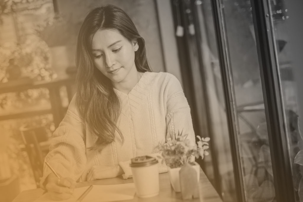 Una ragazza è immersa nella scrittura del suo diario. Il suo viso è consapevole e felice. Sul tavolo ci sono un caffè da asporto e dei fiori.