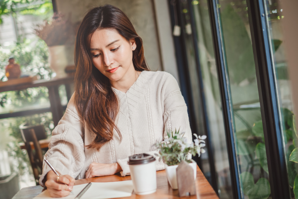 Una ragazza è immersa nella scrittura del suo diario. Il suo viso è consapevole e felice. Sul tavolo ci sono un caffè da asporto e dei fiori.